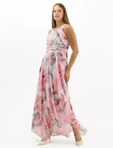 Vestido largo de fiesta con estampado floral en tonos rosa y gris sobre fondo blanco. Presenta cuello halter con abertura tipo lágrima, cintura drapeada y falda amplia con capas asimétricas y bajo en picos. Confeccionado en tejido ligero con forro interior.