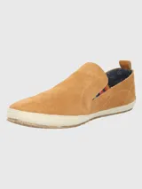 Alpargata Hush Puppies de gamuza color mostaza, con suela de yute trenzado y base de goma. Presenta un detalle elástico lateral con diseño multicolor y forro interior de algodón orgánico.