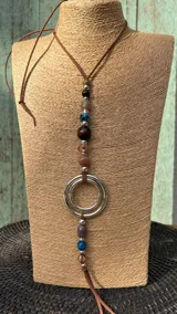 Collar largo con tira de cuerina marrón, decorado con una sección central de cuentas de diferentes tamaños, colores y materiales (madera, piedra y metal), y dos argollas metálicas entrelazadas.