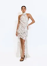 Vestido largo de encaje color marfil, con cuello halter y espalda descubierta. Presenta volados delicados, transparencias y detalles bordados. El diseño es asimétrico, corto en la parte delantera y largo con cola en la parte trasera.