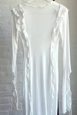 Vestido largo de microtul blanco, con escote en V profundo y mangas largas acampanadas. Presenta aberturas laterales en la falda y cierre con lazos en el frente. Es una prenda transparente.