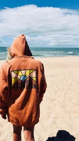 Hoodie color terracota/óxido con capucha y estampado gráfico en la espalda que representa un paisaje montañoso con un sol naciente o poniente y la palabra "MOUNTAIN" en la parte inferior.