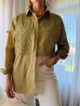 Camisa de manga larga color verde militar, con cuello clásico y cierre frontal de botones. Presenta un canesú bordado con diseño ornamental en la parte frontal superior.