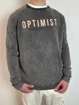 Buzo de cuello redondo con efecto lavado, color gris oscuro. Presenta estampado frontal con la palabra 'OPTIMIST' en letras claras y 'DUXOR COLLECTION' debajo en letras oscuras. Posee puños y ruedo acanalados.