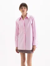 Camisa holgada de poplin blanco, con cuello clásico, mangas largas con puños anchos y cierre frontal con botones. Presenta detalles de apliques metálicos en las puntas del cuello y sutil bordado en el delantero.