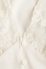 Cropped top de color beige claro, con escote en V pronunciado, mangas cortas abullonadas y cierre frontal con pequeños botones. Presenta detalles de encaje floral en el escote y los puños.