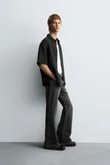 Camisa negra de manga corta con cuello solapa y textura arrugada o plisada. Corte relajado (relaxed fit) con aberturas laterales y cierre frontal de botones.