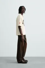 Camiseta de manga corta color crema, corte boxy fit, con cuello redondo. Presenta un estampado frontal en negro con el texto 'VORTEX PULSE' y el número '1974' en un óvalo rojo desgastado. Tiene pequeños detalles gráficos rojos en las mangas.