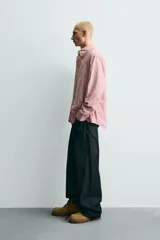 Camisa de corte relajado confeccionada en tejido de algodón, con diseño de rayas verticales en tonos rosados y blancos. Presenta cuello abotonado, cierre frontal de botones, manga larga con puños abotonados y bajo asimétrico.