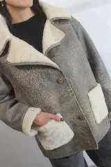 Chaqueta tipo gamulán de cuero genuino en tono gris con efecto desgastado, forrada con piel sintética en color crema. Presenta cuello solapa amplio, cierre frontal con botones y bolsillos de parche con detalle de piel sintética a juego.