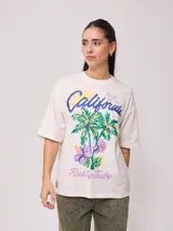 Remera color crudo de corte oversized con estampado de palmeras, flores y texto 'California' y 'Rusty Tribe'.