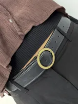 Cinto de cuero negro con textura granulada, costuras decorativas en los bordes y hebilla circular metálica en acabado plateado mate.