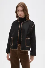 Chaqueta negra de poliéster con cuello a la base, cierre frontal con botones y dos bolsillos delanteros. Presenta detalles de bordado decorativo en color marrón en los bordes, cuello, puños y bolsillos.