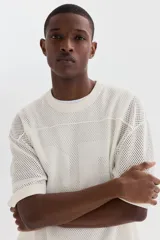 Remera de malla color blanco hueso, de manga corta, con cuello redondo y hombros caídos. Presenta un diseño de tejido perforado con un número grande grabado en el frente.