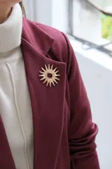 Blazer de corte sastrero en color bordó, con solapas clásicas, bolsillos frontales con solapa y cierre frontal con botones. Presenta una textura sutil en la tela y un broche decorativo en forma de sol en la solapa.