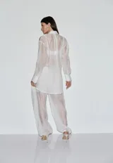 Camisa de organza blanca con transparencias, cuello camisero, mangas largas con puños anchos y detalles de franjas oscuras en los hombros y mangas.