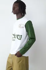 Remera de manga larga con diseño estilo camiseta de fútbol, cuerpo blanco y mangas verdes. Presenta estampados gráficos en el frente con la inscripción 'Victory Legend', el número '39' y un emblema ovalado.