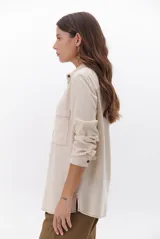 Camisa beige de manga larga, con cuello clásico, botones marrones y dos bolsillos frontales. Presenta costuras decorativas en contraste.