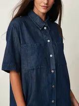 Vestido corto de denim azul oscuro, con cuello camisero, mangas cortas, cierre frontal de botones y dos bolsillos en el pecho.