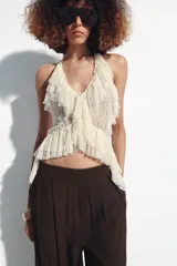 Top de encaje color blanco crudo, con cuello halter, escote en V profundo, volantes en los bordes y bajo asimétrico con lazada en el cuello.