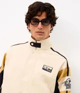 Campera de sarga de algodón marca Blue Steel, con diseño de bloques de color en tonos beige, negro y mostaza. Presenta cuello alto, cierre frontal con botones a presión y solapa, bolsillos laterales y detalles de parches bordados con temática outdoor en el pecho y la manga.