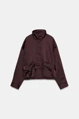 Cazadora satinada de color negro, con cuello subido, manga larga y bolsillos frontales de plastrón con cordones decorativos. Cuenta con bajo elástico y cierre frontal con cremallera y botones a presión oculta.