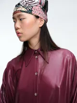 Camisa de corte oversize confeccionada en tejido jersey con acabado brillante. Presenta cuello en punta, cierre frontal con botones, mangas largas con puños anchos y tajos laterales en el ruedo.