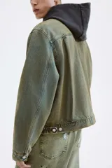 Campera de denim de algodón rígido, color verde azulado desgastado, con cuello, cierre frontal de botones metálicos y dos bolsillos superiores con solapa y botón. Presenta puños largos abotonados y trabillas ajustables con botones en los laterales.