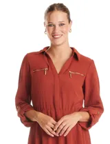 Vestido corto color terracota, confeccionado en crepe, con cuello camisero, escote en V y cierre frontal parcial. Presenta bolsillos decorativos con cierres metálicos en el pecho, mangas largas regulables con presillas y cintura elastizada.