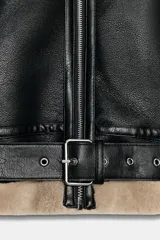 Cazadora de estilo aviador con exterior de cuero sintético negro y forro interior de piel sintética en tono beige. Presenta cuello solapa, cierre frontal con cremallera, bolsillos laterales con cremallera, trabillas en los puños y cinturón ajustable en el bajo con hebilla metálica.