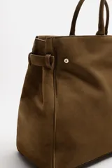 Bolso de mano extragrande color marrón oscuro, estilo tote o satchel, fabricado en piel. Presenta dos asas de mano y un detalle de cinta ajustable con cierre de solapa en la parte frontal superior.