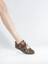 Sneaker de perfil bajo Schutz con estampado animal print tipo cebra en textura de pelo. Presenta cordones negros y blancos, con un detalle de parche triangular en color rojo en la lengüeta y suela plana de goma.