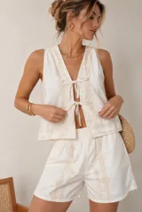 Conjunto de dos piezas color blanco, compuesto por una blusa sin mangas con escote en V y cierre de lazos frontales, y un short a juego. Ambas prendas cuentan con delicados detalles bordados florales en tono beige.