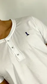 Remera tipo Henley de manga corta en color blanco, con cuello redondo y tapeta frontal con tres botones. Presenta un pequeño logo bordado en el lado izquierdo del pecho.