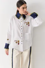 Camisa blanca de corte holgado con cuello y puños tejidos en crochet azul con borde verde. Presenta parches bordados decorativos en el frente y las mangas, cierre frontal con botones y un bolsillo de parche en el pecho.