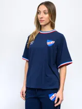 Remera blanca de manga corta con cuello redondo y puños ribeteados en rojo, blanco y azul. Presenta el escudo del Club Nacional de Football bordado en el pecho izquierdo y el logo de Umbro bordado en el pecho derecho.