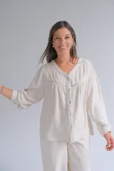 Blusa de lino color beige, con cuello en V, cierre frontal con botones y detalle de alforzas y encaje en el pecho. Presenta mangas largas con puños sencillos y un corte holgado.