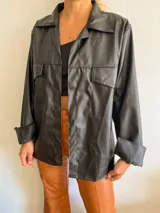 Camisa negra de eco cuero con cuello clásico, solapas y cierre frontal con botones a presión. Tiene dos bolsillos de parche en el pecho y mangas largas con puños abotonados.