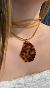 Gargantilla tipo choker con cordón de gamuza color tostado que se envuelve varias veces al cuello, con un dije grande de resina con estampado tipo carey en tonos marrones.