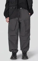 Pantalón cargo oversize de color gris, confeccionado en tejido técnico ripstop. Cuenta con cintura elástica con presillas, cierre frontal con broche técnico, múltiples bolsillos amplios y sistema de ajuste con broches snap en los tobillos.