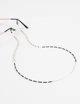 Correa para lentes confeccionada con canutillos de vidrio en tonos blanco, negro y beige, con terminaciones metálicas doradas y sujetadores de goma negros.