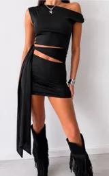 Vestido corto negro de diseño asimétrico con escote de un solo hombro y detalle de cut-out en la cintura con una faja lateral larga que cae hacia un costado.