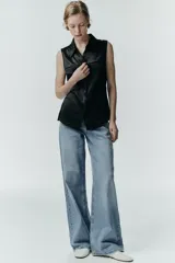 Camisa sin mangas de tejido satinado, con cuello solapa, cierre frontal con botones ocultos y bolsillos de plastrón con solapa en el delantero. Presenta un bajo asimétrico y corte recto.