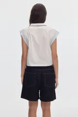 Camisa cropped sin mangas de algodón, con diseño colorblock en tonos blanco y celeste, cuello camisero con ribete negro, cierre frontal de botones y bolsillos en el pecho.