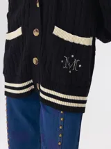 Cardigan de punto trenzado en color negro con detalles en color crema en los bolsillos, puños y ruedo. Presenta un diseño oversize, cierre frontal con botones y bordado decorativo en uno de los bolsillos delanteros.