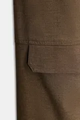 Pantalón de tiro medio color marrón oscuro, estilo cargo, con cintura acabada en vuelta asimétrica, pliegues delanteros y bolsillos laterales y con solapa en las piernas.