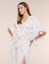 Blusa blanca de silueta holgada con sutil trama en hilo metalizado lurex, escote en V con pechera tejida en crochet y apliques de borlas en el contorno del cuello y las mangas.