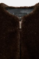 Chaqueta bomber de cuello redondo y manga larga, confeccionada en tejido de pelo sintético color marrón oscuro. Presenta cierre frontal con cremallera, bolsillos laterales y acabados elásticos en el ruedo y puños.