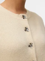 Cardigan de punto color beige con cuello redondo y mangas largas. Presenta un cierre frontal con botones metálicos decorativos en forma de flor plateada.