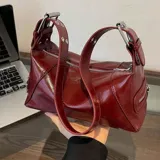 Cartera tipo baguette de cuero sintético color borgoña, con doble cierre superior y correa corta de hombro con hebillas metálicas.